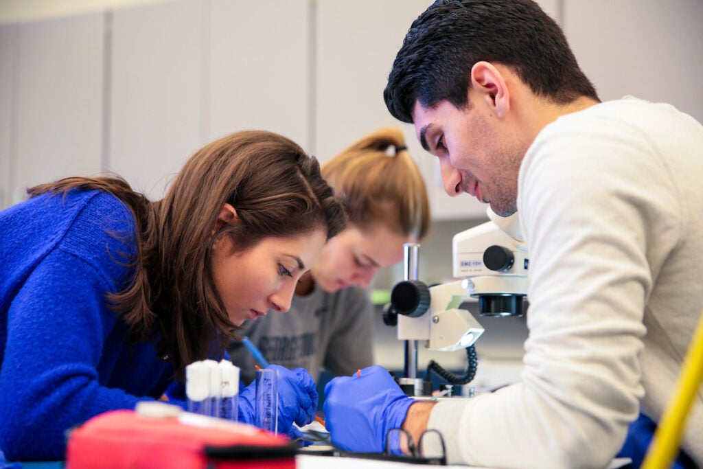 Undergraduate students in a lab writing down notes. They are wearing PPE and microscopes can be seen in the background.