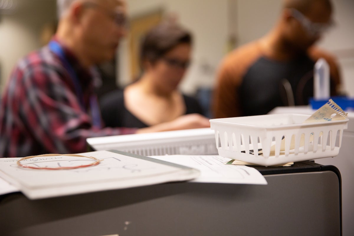 Notebooks, papers, and trays on top of a desk. Blurred figured of students and faculty are seen in the background.